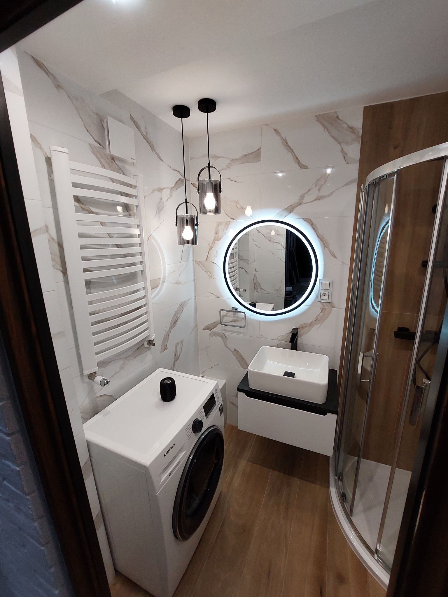 Nowoczesna łazienka z marmurowymi płytkami, okrągłym lustrem z podświetleniem LED, wiszącą umywalką, grzejnikiem drabinkowym i pralką Samsung.