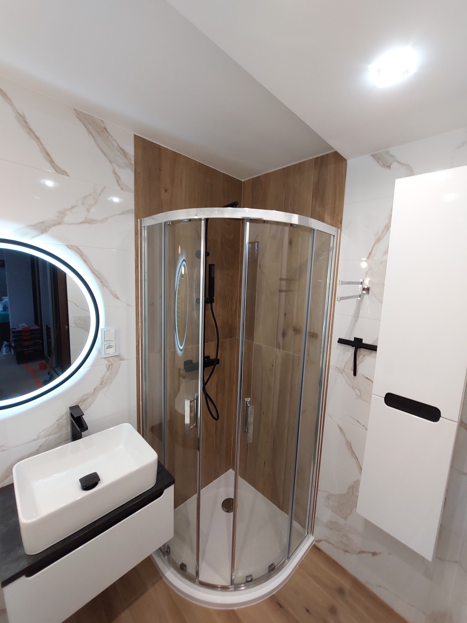Nowoczesna łazienka z kabiną prysznicową narożną, umywalką nablatową na szafce z ciemnym blatem, okrągłym lustrem z podświetleniem LED i płytkami imitującymi marmur z delikatnym, złotym użyleniem.