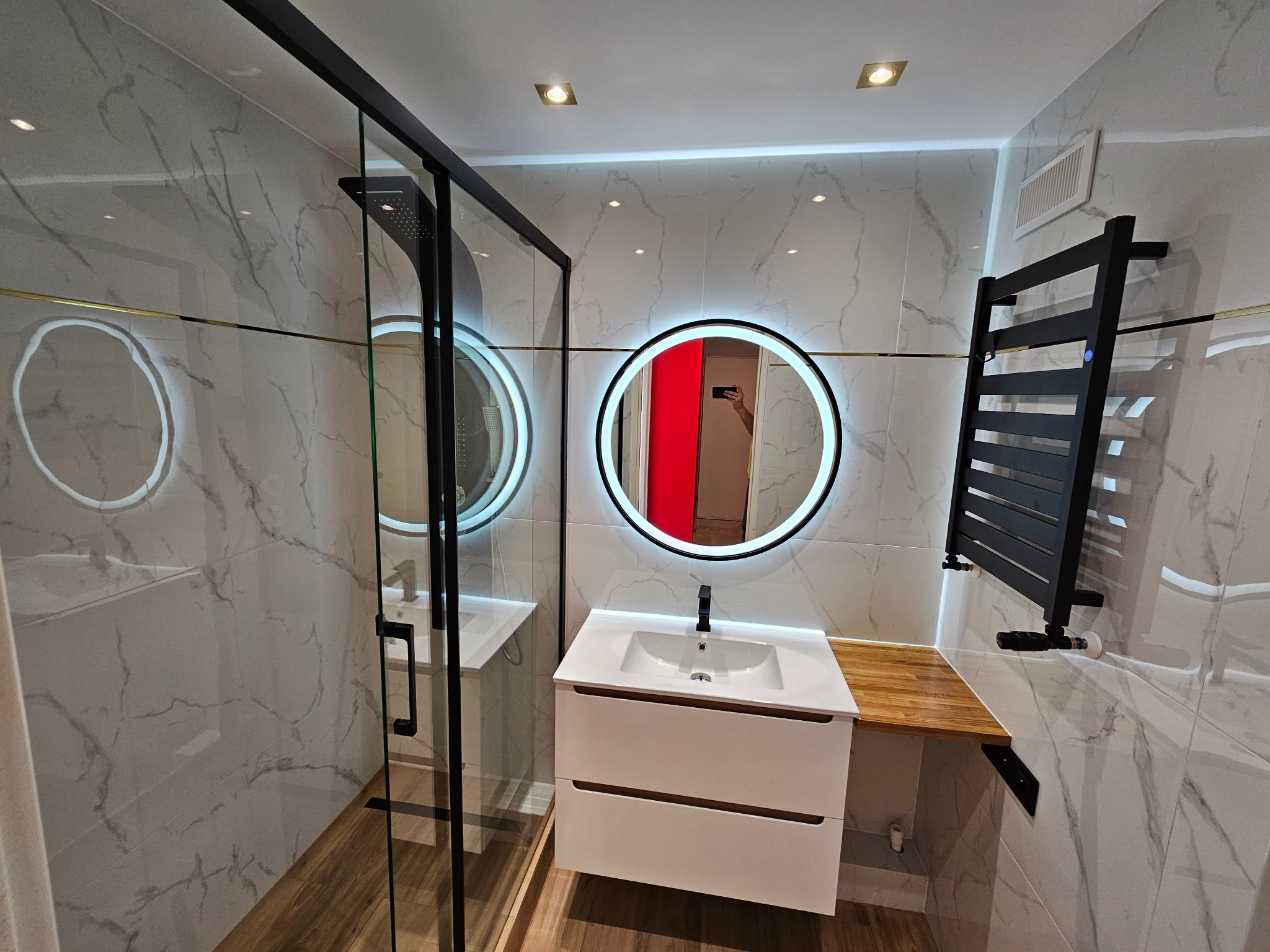 Nowoczesna łazienka z kabiną prysznicową typu walk-in, okrągłym lustrem z podświetleniem LED, białą szafką pod umywalką i czarnym grzejnikiem drabinkowym na tle marmurowych płytek.