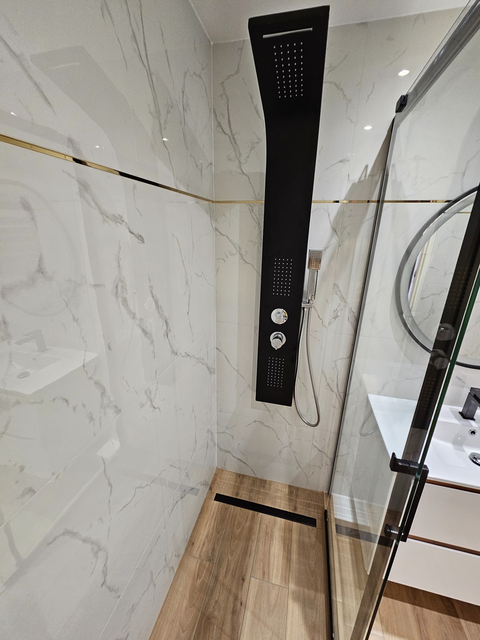 Nowoczesna kabina prysznicowa z czarnym panelem natryskowym, marmurowymi płytkami ściennymi z delikatnym złotym akcentem i drewnopodobną podłogą, widok na umywalkę z okrągłym lustrem.