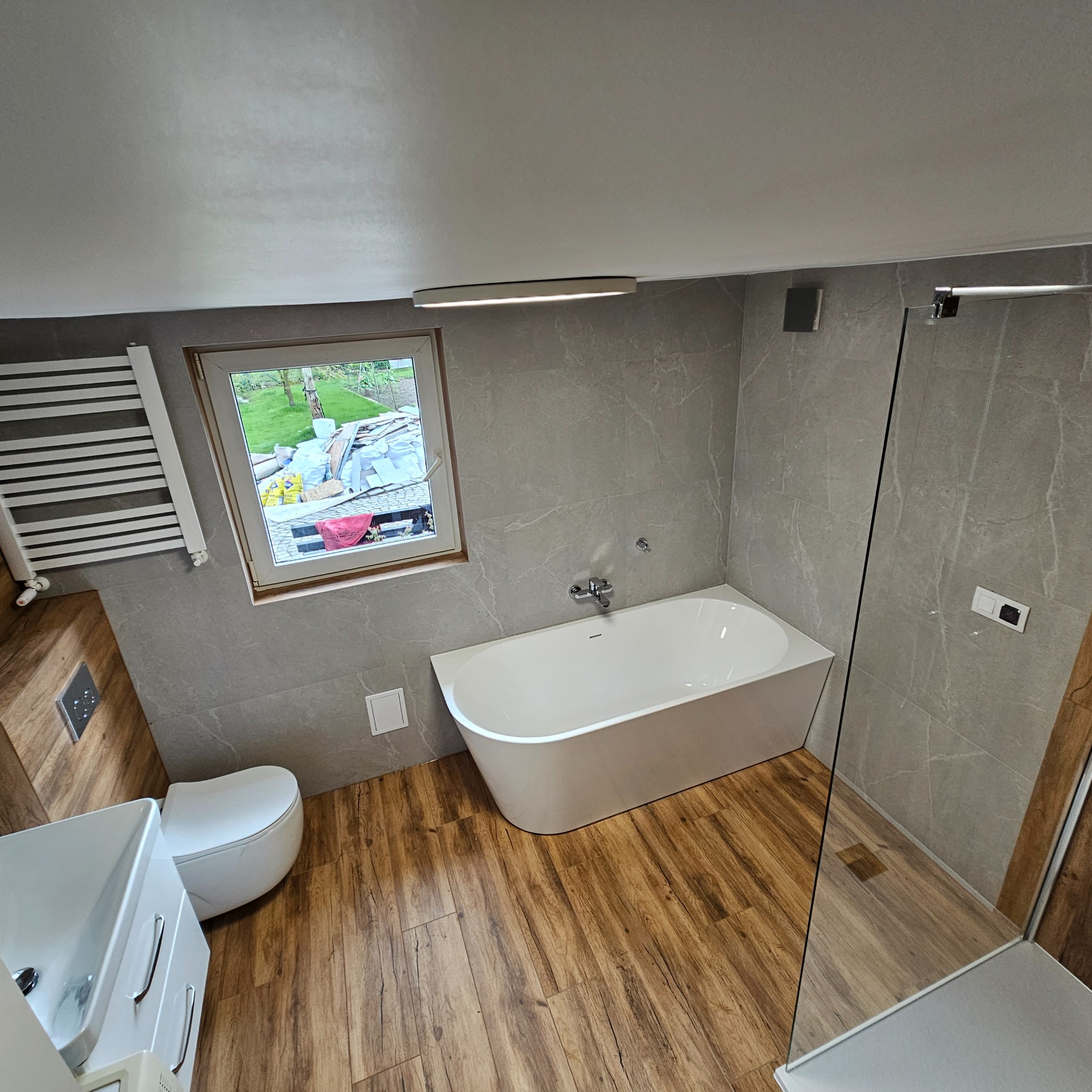 Nowoczesna łazienka z drewnopodobną podłogą, szarymi płytkami imitującymi marmur na ścianach, białą wanną wolnostojącą i przeszkloną kabiną prysznicową. Za oknem widoczny bałagan budowlany.