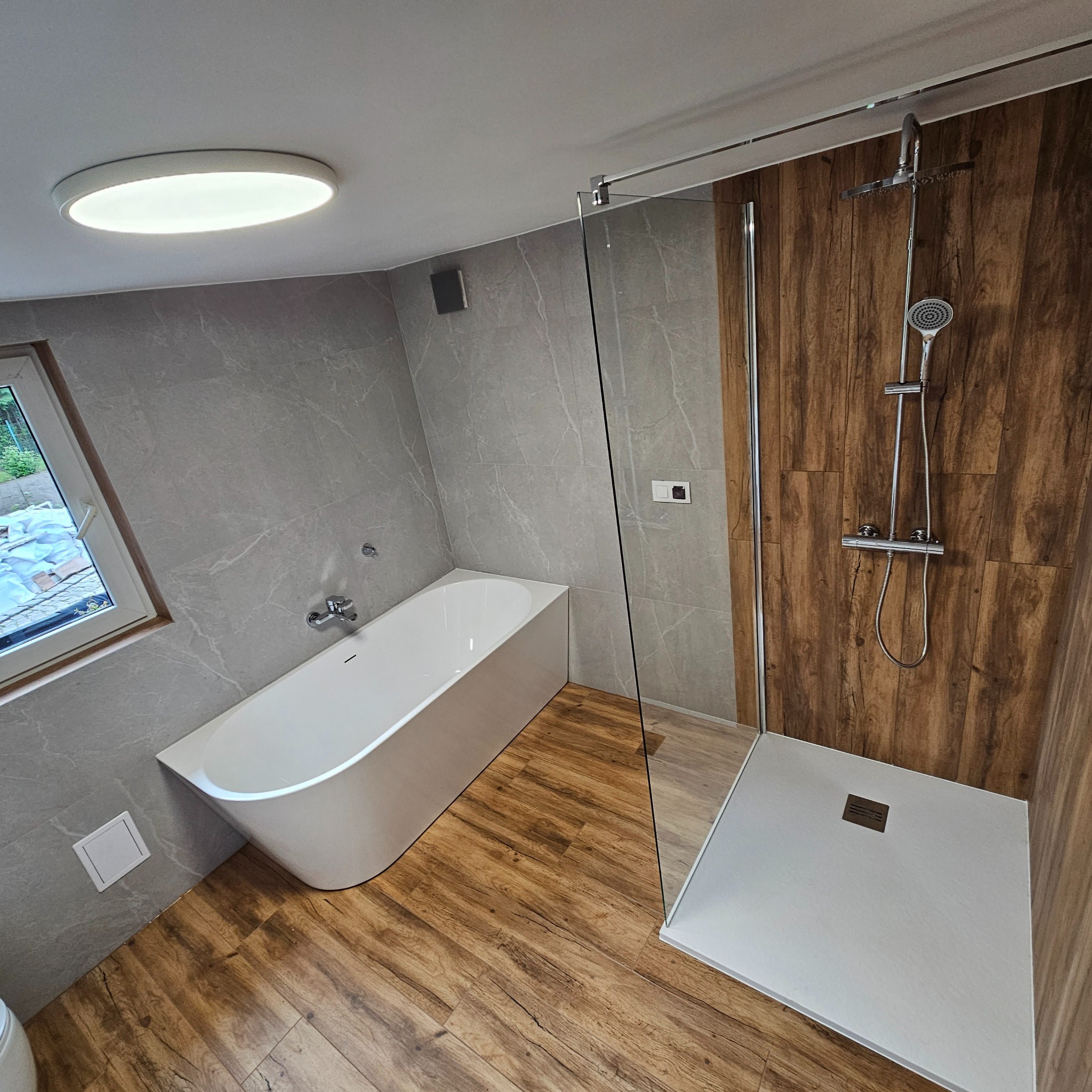 Nowoczesna łazienka z wolnostojącą wanną, szarą marmurkową okładziną ścian, drewnopodobną podłogą i kabiną prysznicową ze szklaną ścianką. Widoczne okno z widokiem na zewnątrz.