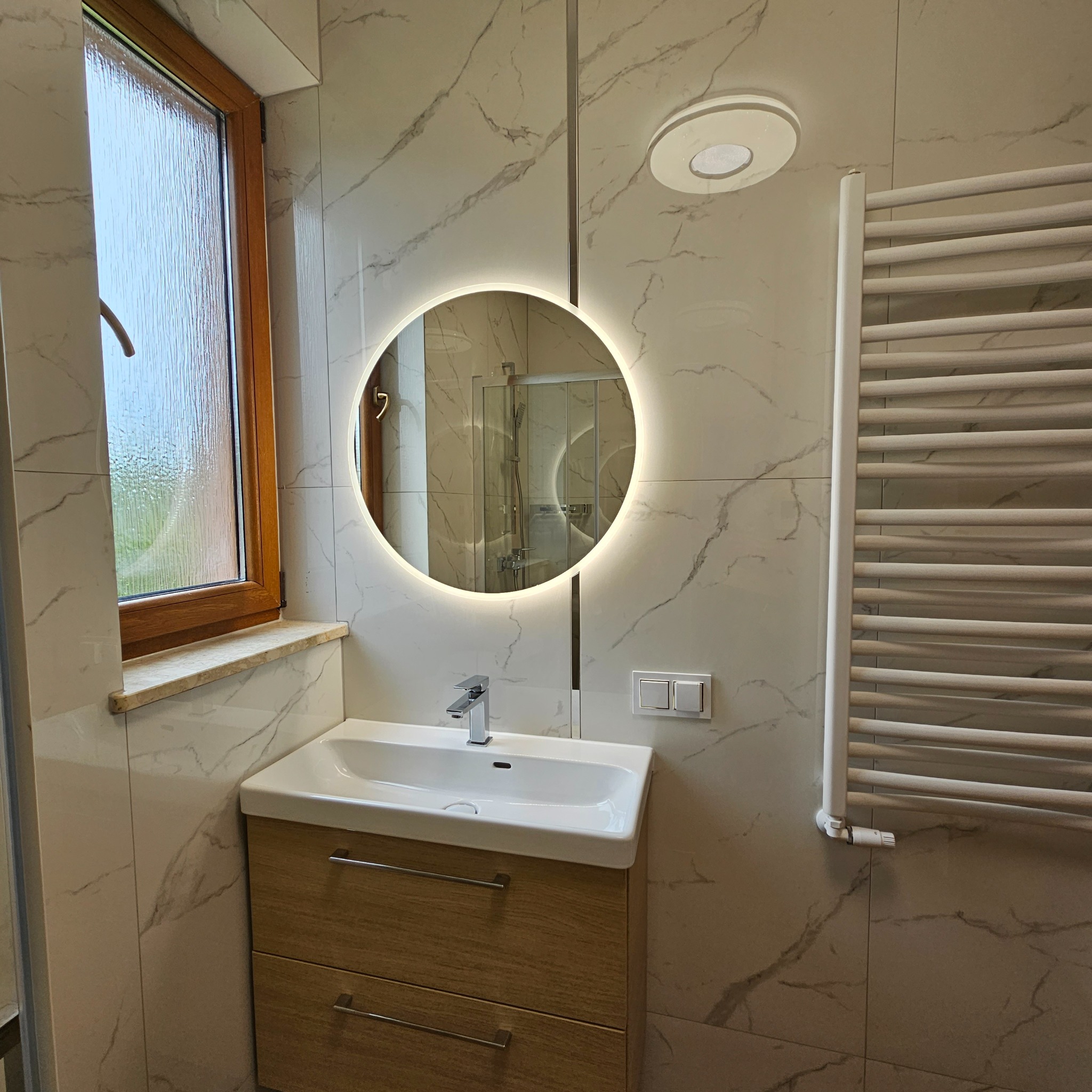 Wykończona łazienka z okrągłym lustrem z podświetleniem LED, umywalką nablatową z szafką z drewnopodobnym fornirem, grzejnikiem drabinkowym i marmurowymi płytkami ściennymi.