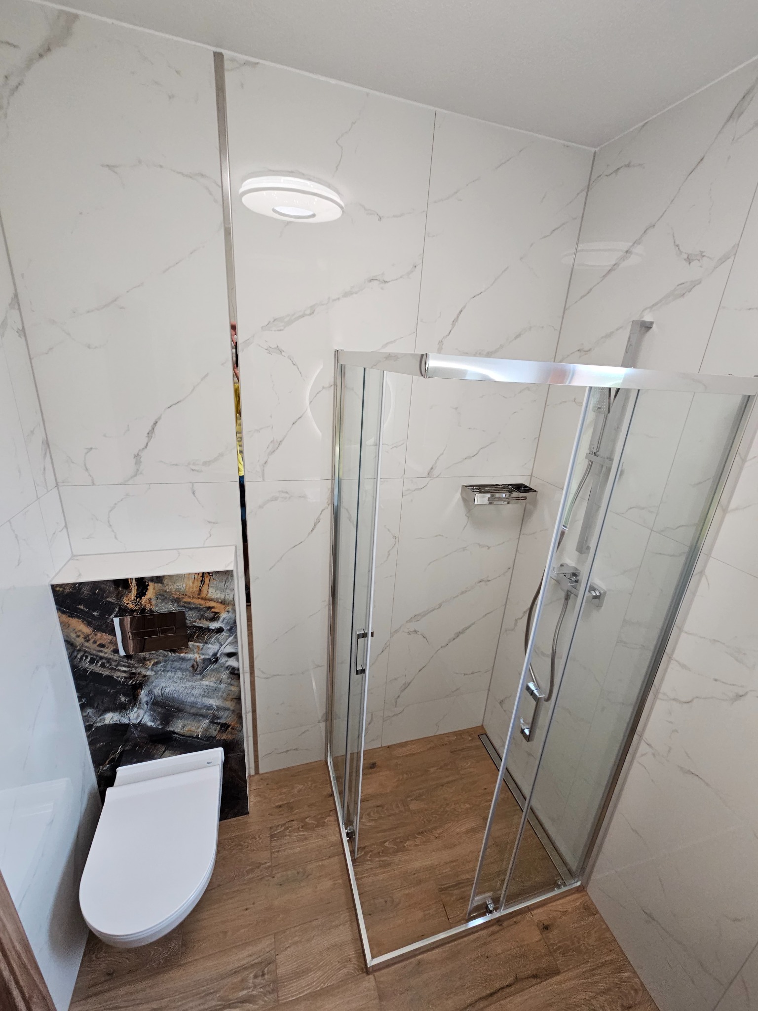 Nowoczesna łazienka z kabiną prysznicową ze srebrnymi profilami, podwieszaną toaletą z czarną obudową imitującą kamień oraz płytkami imitującymi biały marmur na ścianach i drewnopodobną podłogą.