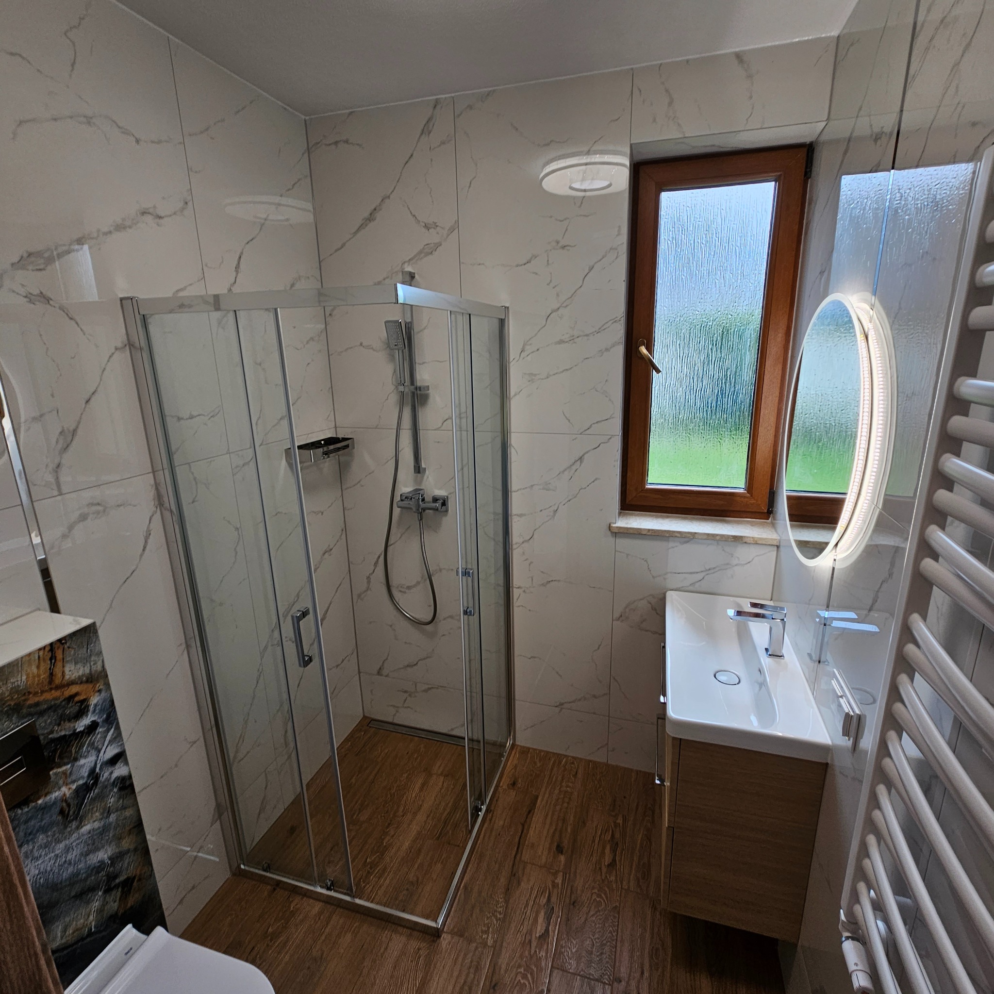 Nowoczesna łazienka z kabiną prysznicową, umywalką z szafką z drewnopodobnym dekorem i podświetlanym lustrem owalnym, wykończona płytkami imitującymi marmur i drewno.