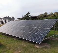 Panel słoneczny ustawiony na trawiastym terenie przed domem jednorodzinnym. Widoczna konstrukcja wsporcza paneli i przewody. Ujęcie z niskiej perspektywy.