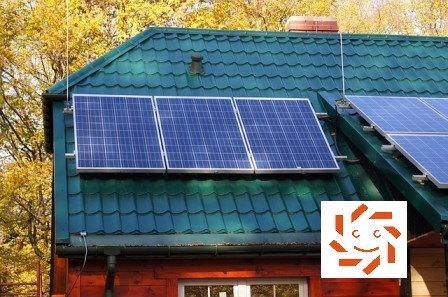 Instalacja fotowoltaiczna na zielonym, metalowym dachu domu z drewnianą elewacją, widoczna antena i jesienne drzewa w tle.