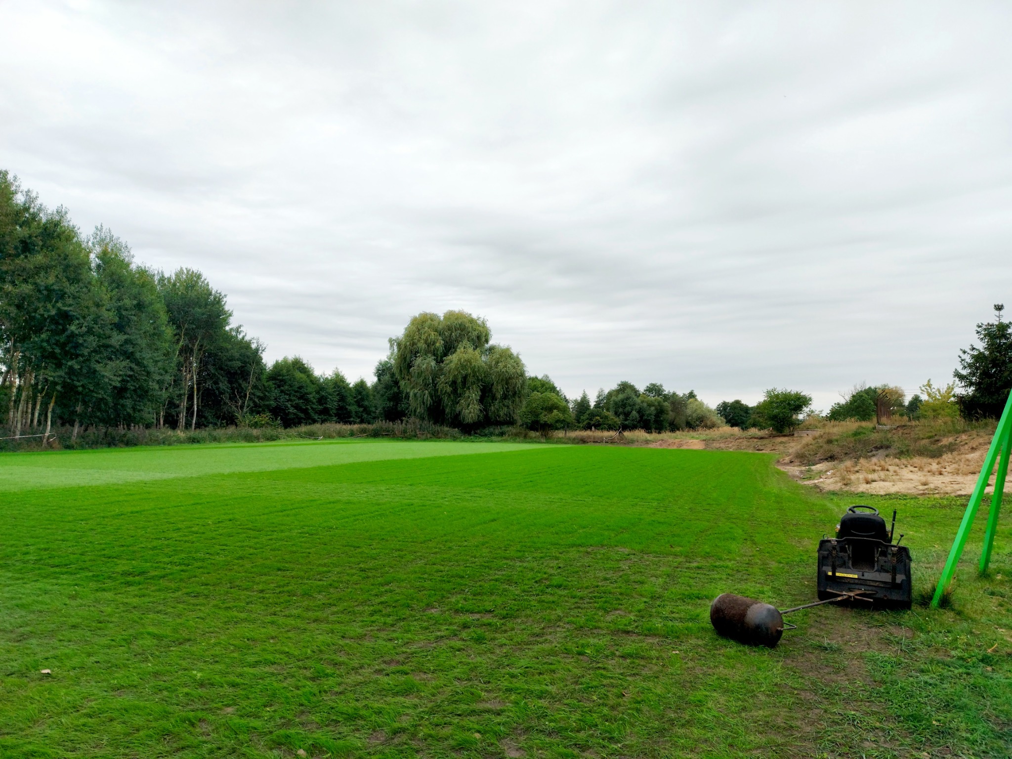Rozległy trawnik z widocznymi śladami koszenia, z tyłu las, a w prawym dolnym rogu kosiarka traktorowa z wałem.