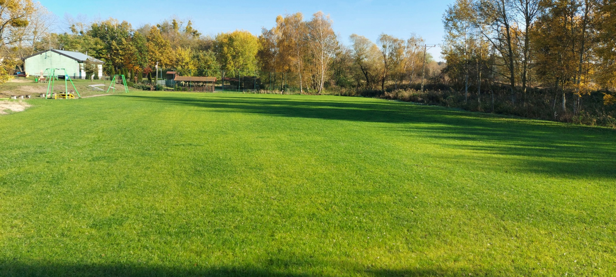 Rozległy trawnik z intensywną zielenią, częściowo zacieniony, z widocznymi drzewami w tle o jesiennych barwach i elementami architektury ogrodowej oraz placu zabaw w oddali.