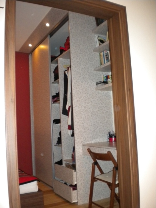 Wnętrze garderoby z przesuwnymi drzwiami, półkami na książki i składanym biurkiem z krzesłem, wykończone tapetą we wzory.