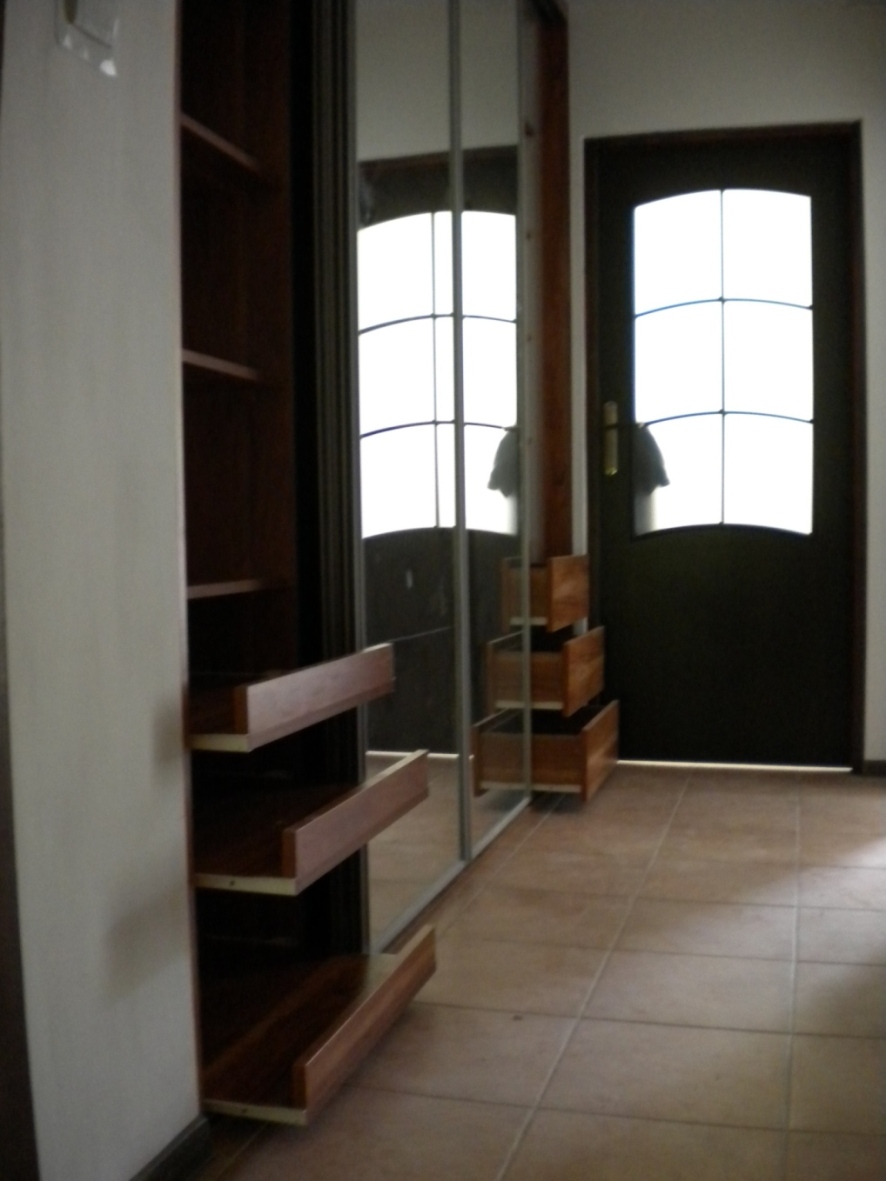 Wnętrze z szafą wnękową z przesuwnymi drzwiami lustrzanymi i otwartymi półkami z drewna w kolorze ciemnego brązu; widoczna część wnętrza z brązowymi płytkami podłogowymi i ciemnymi drzwiami z szybą.