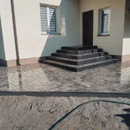 Nowo ułożona, szara kostka brukowa przed domem, z widocznymi schodami wejściowymi i fragmentem węża ogrodowego na piasku.