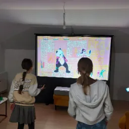 Grupa dzieci grająca w grę taneczną wyświetlaną na projektorze w domowym zaciszu, widoczny ekran projekcyjny, projektor na żółtym stoliku, półki z modelami samolotów w tle.