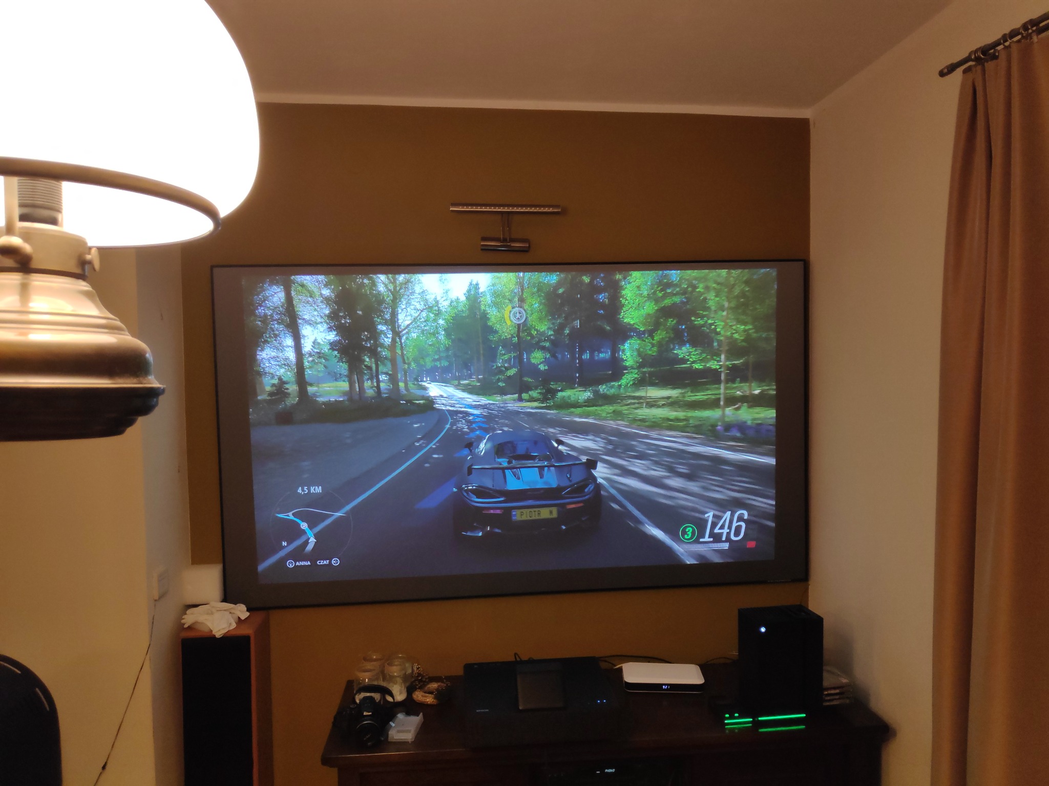 Duży ekran telewizora wyświetlający grę wyścigową, na dole konsola Xbox Series X z zielonym podświetleniem, na szafce dodatkowe akcesoria.