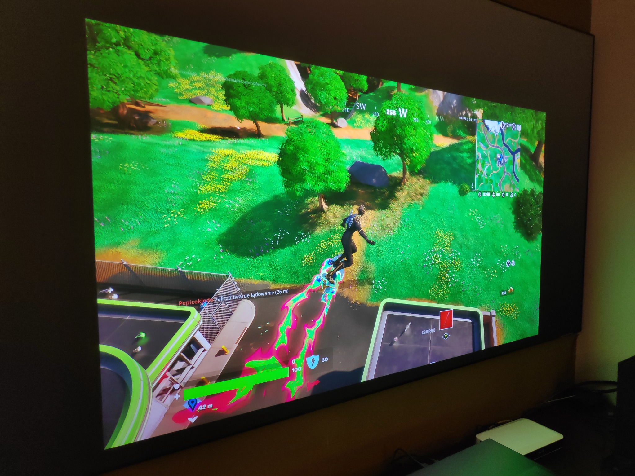 Ekran telewizora wyświetlający rozgrywkę w grę Fortnite, z widoczną postacią w trakcie lotu nad terenem z zieloną roślinnością i elementami budowli, w domu w Wieliczce.