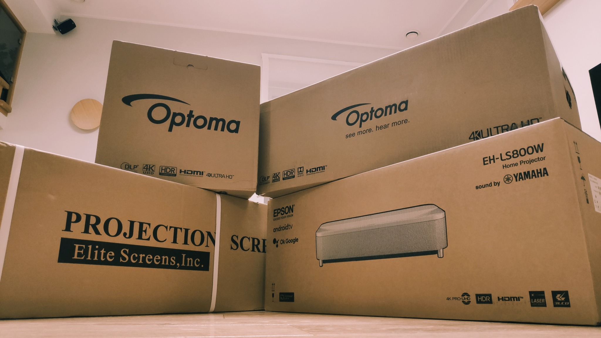 Stos kartonowych pudełek z nadrukami marek Optoma, Epson i Elite Screens, zawierających projektory i ekrany projekcyjne, leżących na podłodze.