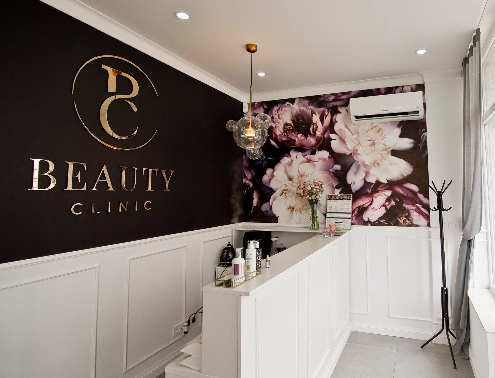 Recepcja w salonie kosmetycznym z klimatyzatorem ściennym na ścianie z motywem kwiatowym, widoczne logo 'Beauty Clinic' w złotym kolorze na czarnej ścianie.