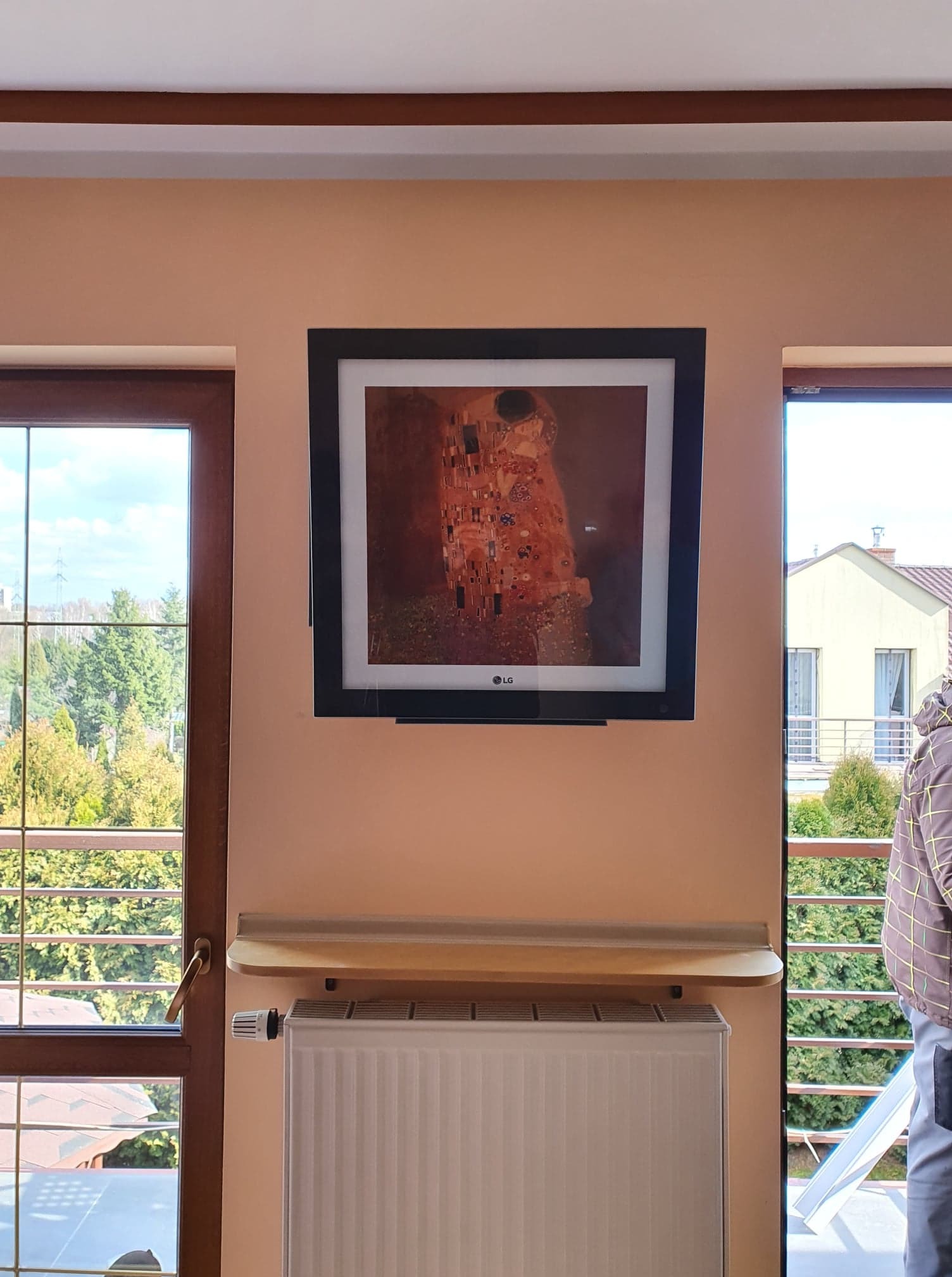 Grzejnik panelowy z termostatem w Chorzowie, zamontowany pod półką, nad nim telewizor LG z obrazem Klimta, widok z okna na zieleń.