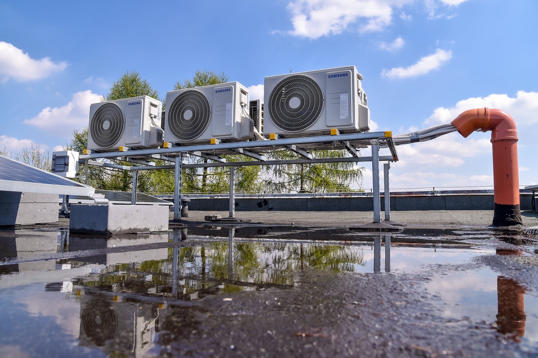Trzy jednostki zewnętrzne klimatyzacji Samsung zamontowane na metalowej konstrukcji na dachu, z odbiciem w kałużach po deszczu i widocznym kominem wentylacyjnym