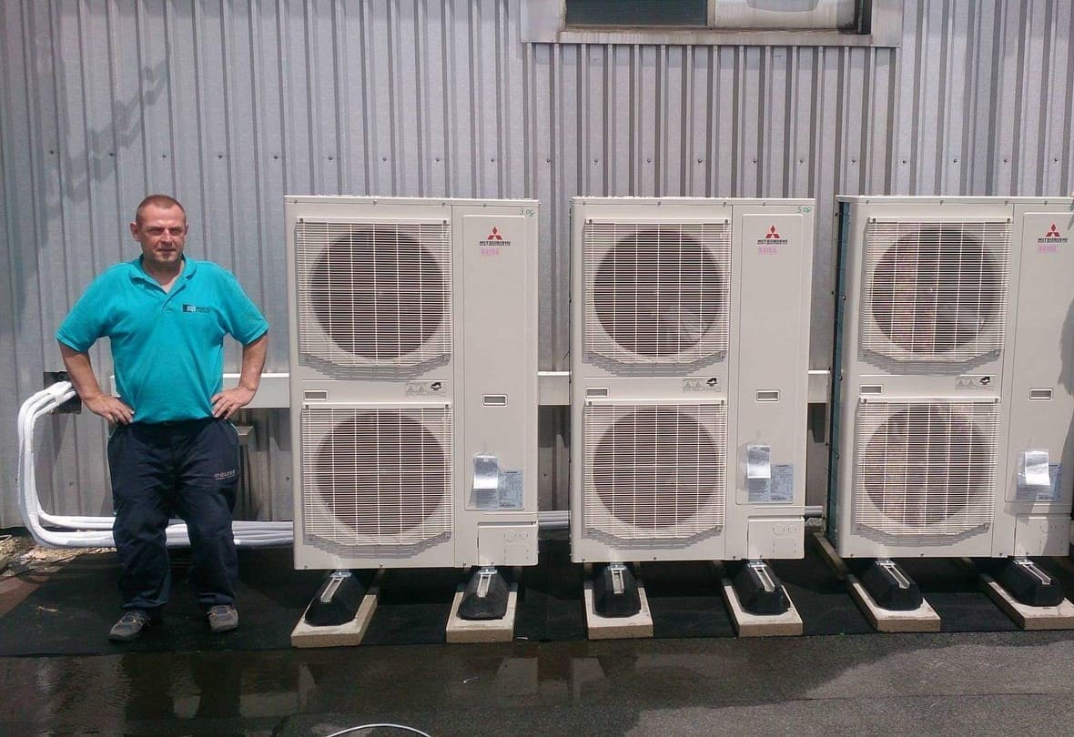 Instalator prezentuje rząd trzech zewnętrznych jednostek klimatyzacji Mitsubishi na betonowych podstawach, zamontowanych na dachu budynku przemysłowego z blachy falistej.