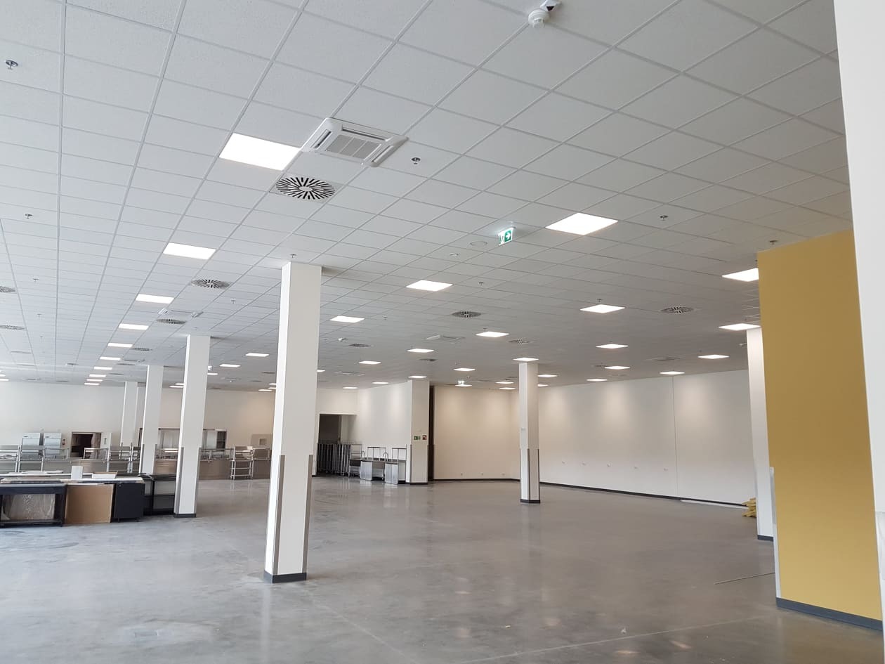 Wnętrze budynku komercyjnego z zainstalowaną wentylacją mechaniczną: widoczne anemostaty sufitowe, kratki wentylacyjne i klimakonwektor kasetonowy w suficie podwieszanym.