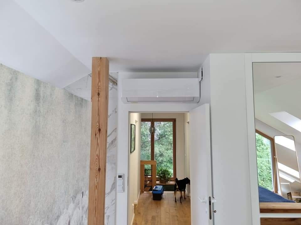 Biały klimatyzator ścienny zamontowany nad drzwiami w jasnym wnętrzu domu z drewnianą kolumną i widokiem na zieleń przez okno, z psem w tle.