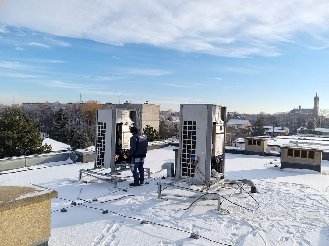 Pracownik w ciemnej kurtce serwisuje dwie szare jednostki klimatyzacyjne na płaskim, ośnieżonym dachu budynku z widokiem na miasto w tle, niebieskie niebo z chmurami.