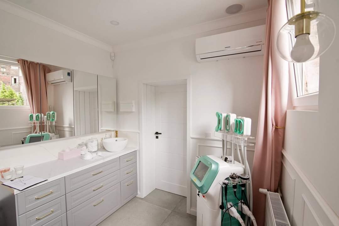 Wnętrze gabinetu kosmetycznego z klimatyzatorem Samsung na ścianie, obok okna z różową zasłoną i specjalistycznym sprzętem kosmetycznym z ekranem i różnymi końcówkami.