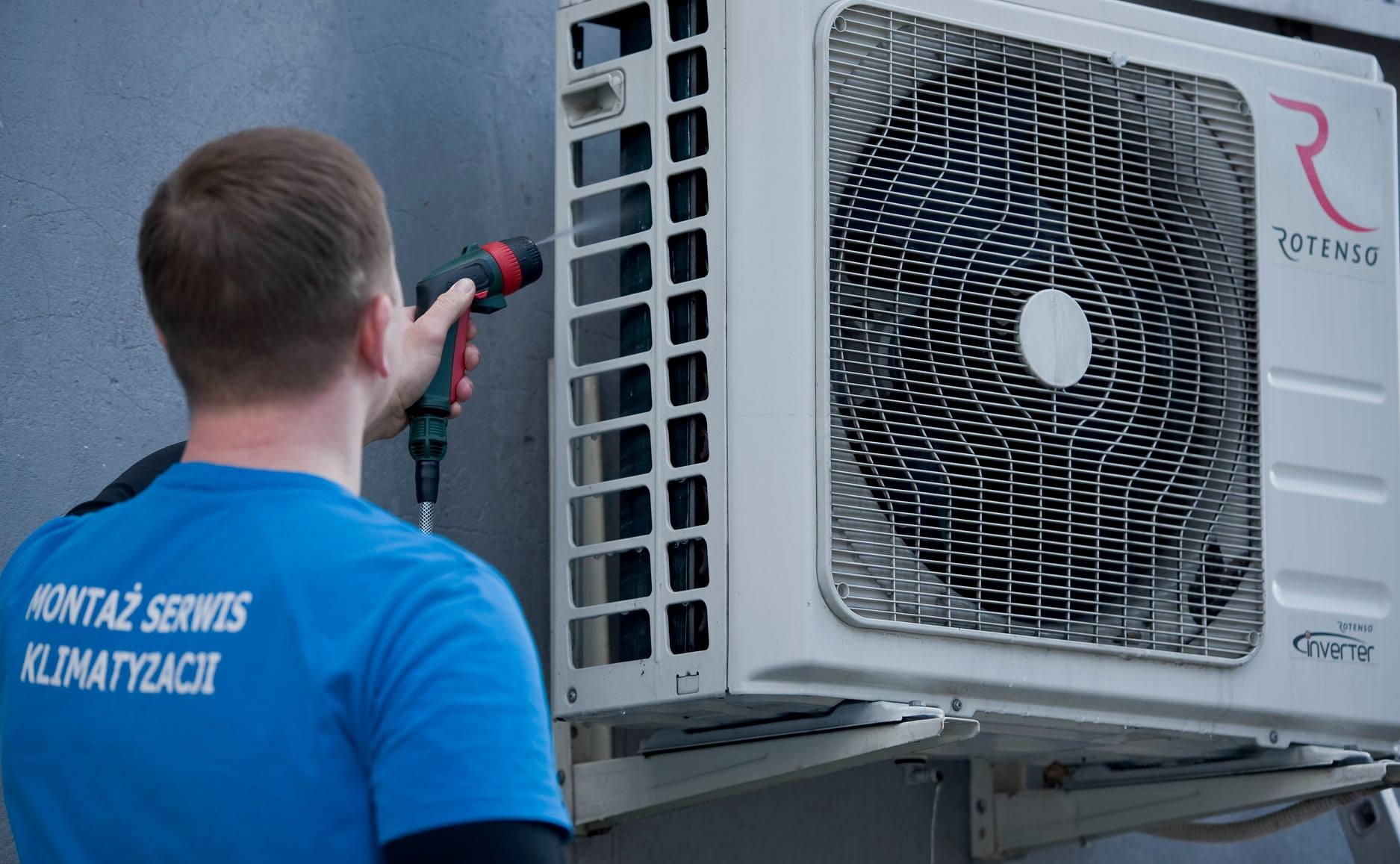 Pracownik w niebieskiej koszulce z napisem 'Montaż Serwis Klimatyzacji' czyści zewnętrzną jednostkę klimatyzatora Rotenso za pomocą węża z pistoletem natryskowym.