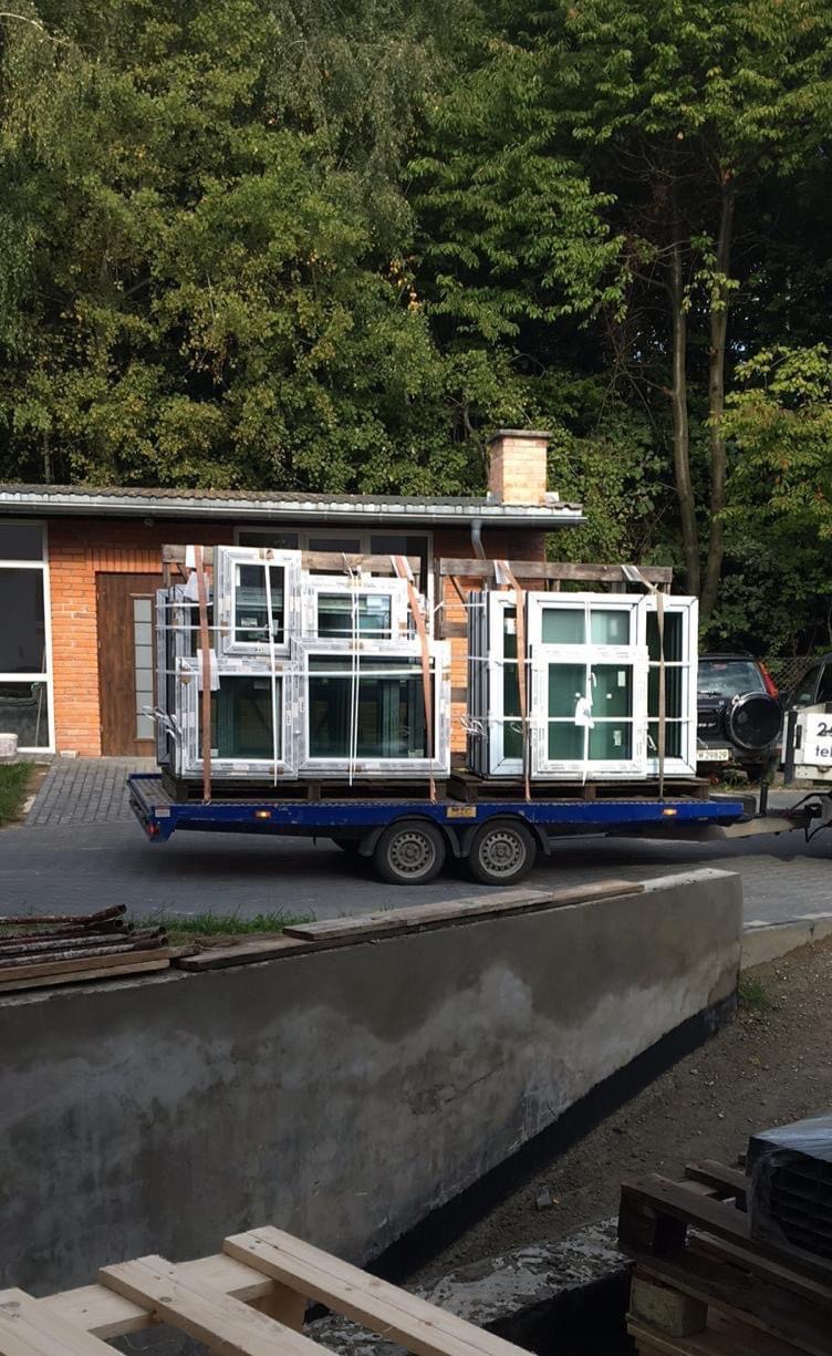 Białe okna PCV zabezpieczone taśmami transportowymi na niebieskiej przyczepie, w tle budynek z cegły i zieleń.