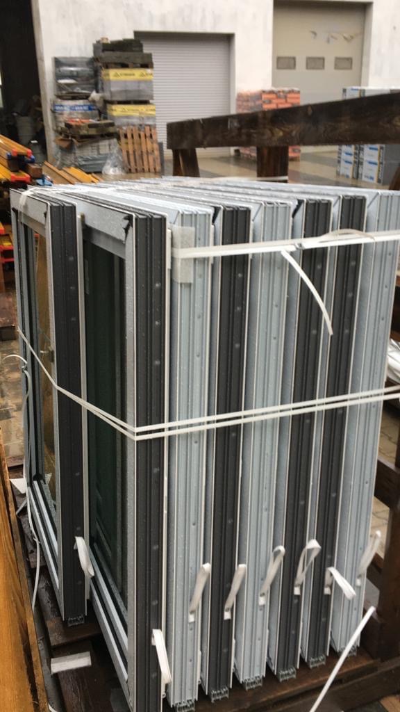 Paczka nowych, szarych ram okiennych z czarnymi uszczelkami, spiętych białym sznurkiem, składowana w magazynie z materiałami budowlanymi.