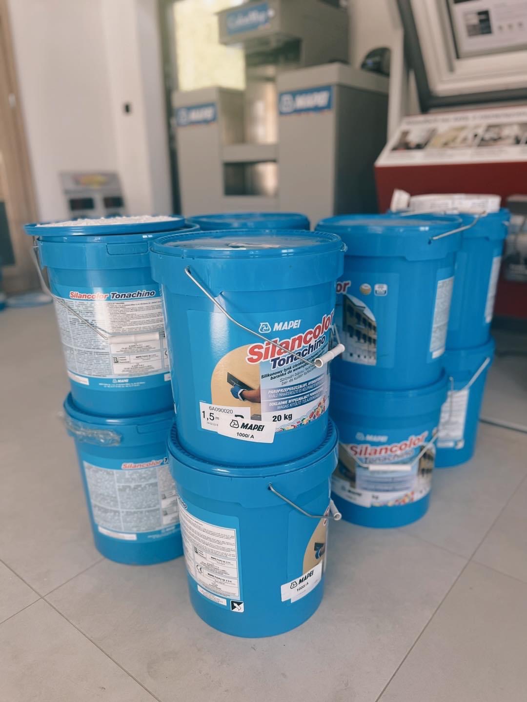Stos niebieskich wiader z silikonowym tynkiem barankiem Silancolor Tonachino firmy Mapei, ustawionych na szarej podłodze w pomieszczeniu.