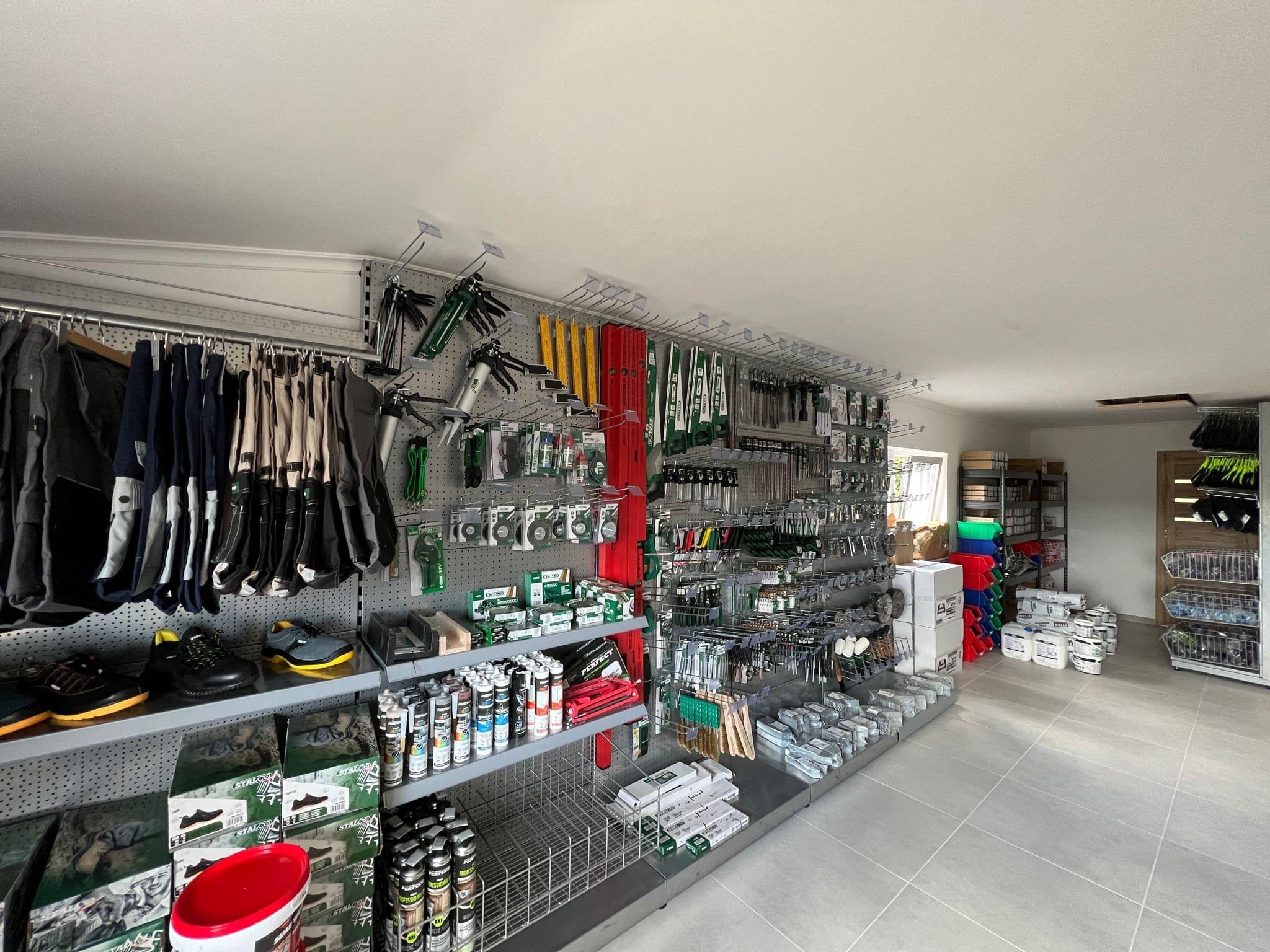 Sklep z narzędziami budowlanymi i odzieżą roboczą: ekspozycja z różnorodnym asortymentem, w tym pistolety do silikonu, poziomice, pędzle, farby, kleje, obuwie i ubrania robocze.