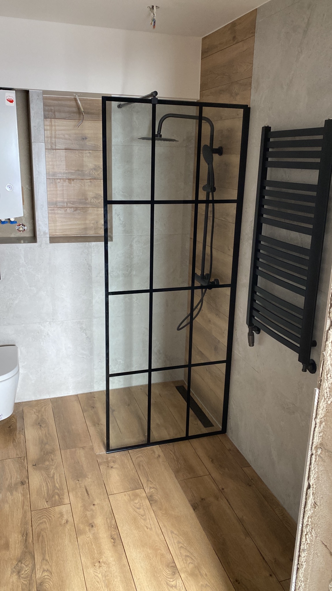 Nowoczesna kabina prysznicowa z czarną ramą i deszczownicą, zamontowana w łazience z panelami imitującymi drewno i grzejnikiem drabinkowym w kolorze czarnym.