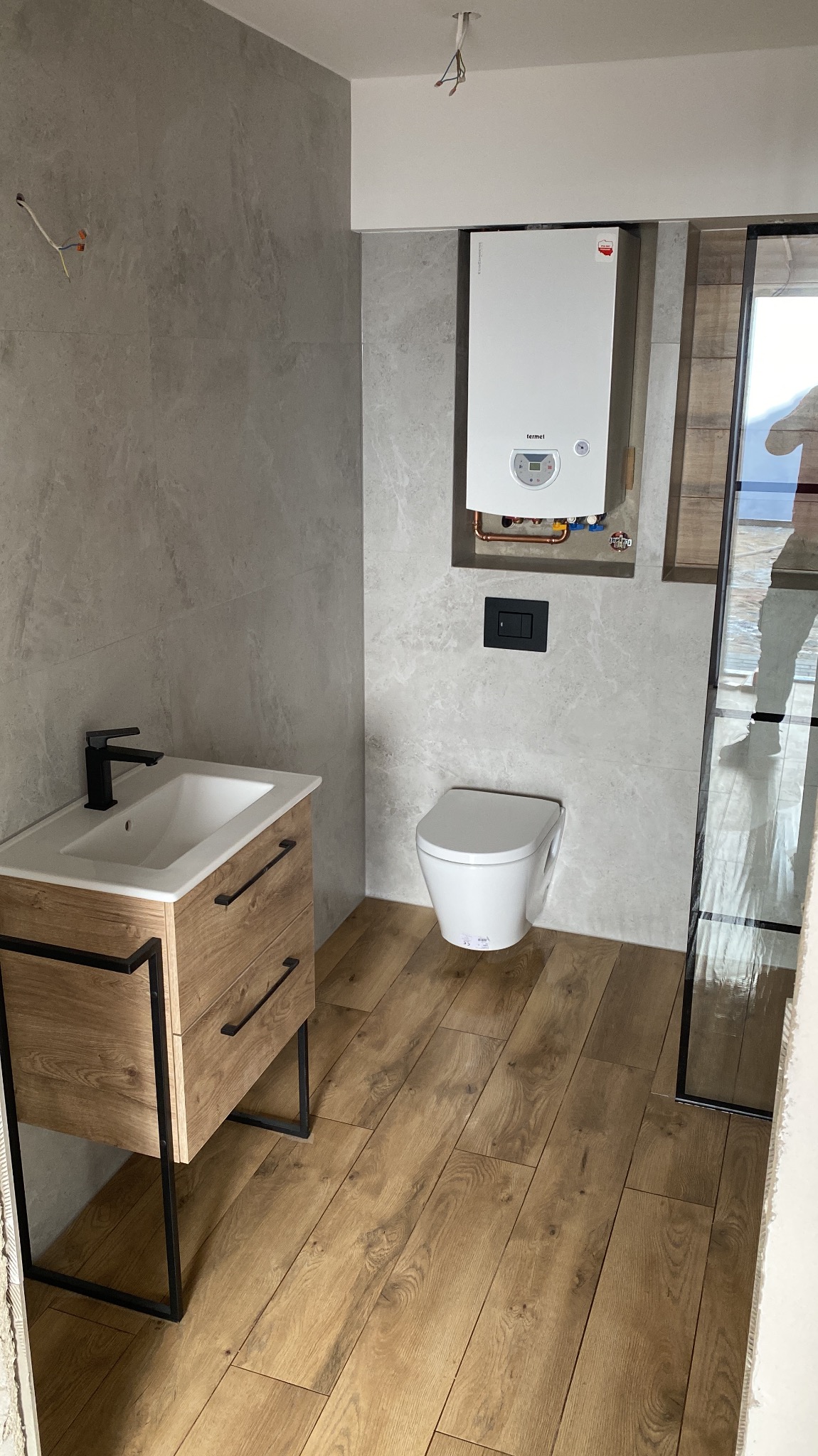 Nowoczesna łazienka z umywalką na szafce z drewnianymi frontami, wiszącą toaletą, szarymi płytkami imitującymi kamień na ścianach i panelami podłogowymi imitującymi drewno, z widocznym piecem...