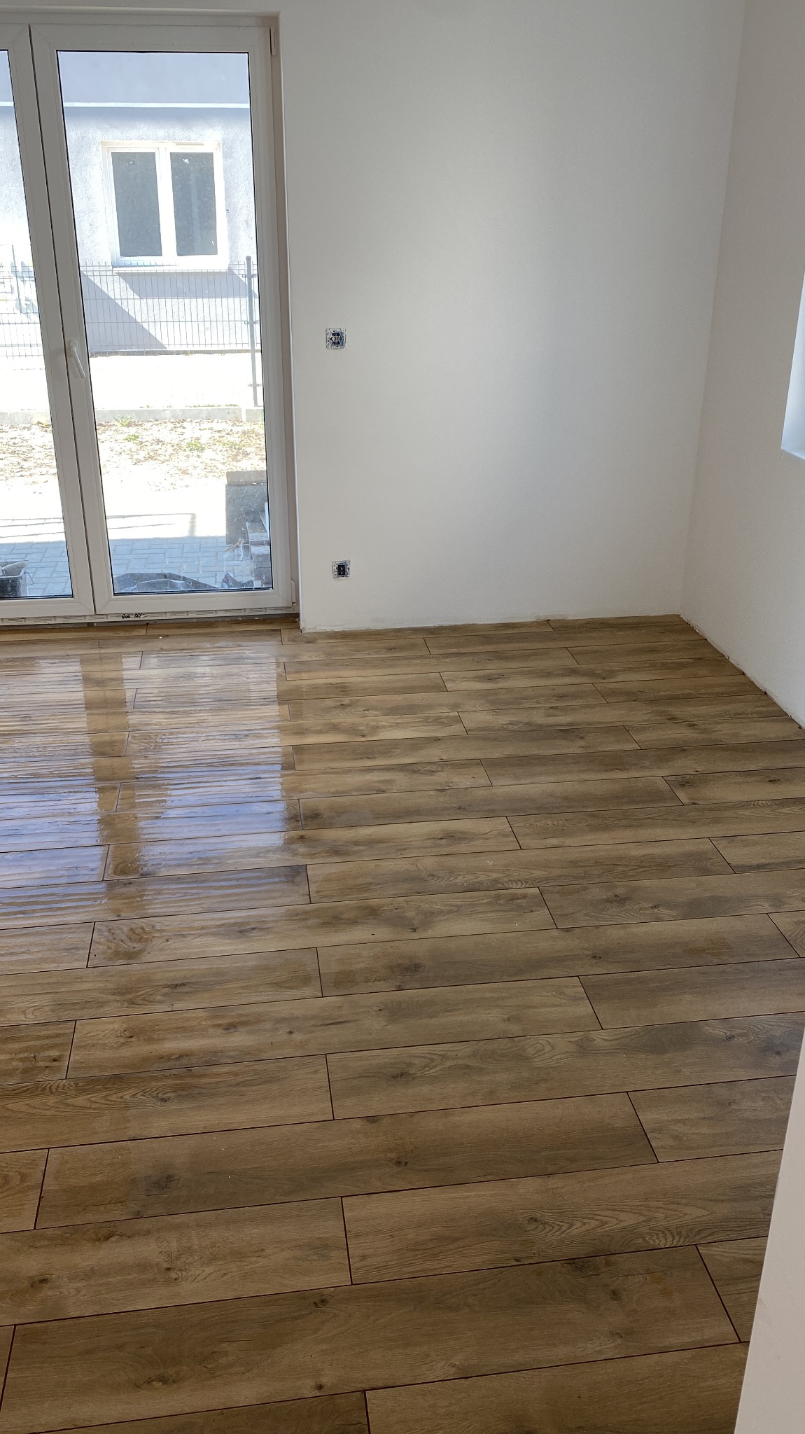 Ułożona podłoga z płytek imitujących drewno w nowym pomieszczeniu z dużym oknem balkonowym i widocznymi gniazdkami elektrycznymi na białej ścianie.