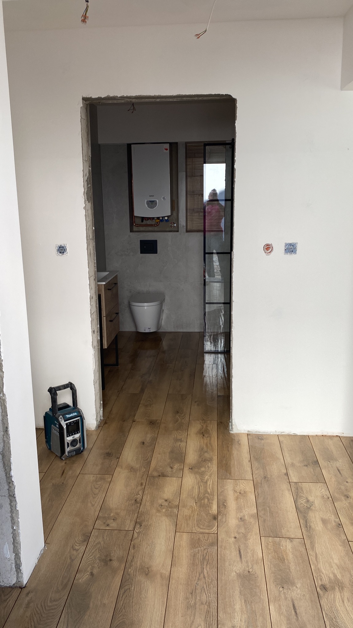 Wnętrze łazienki w trakcie prac wykończeniowych: widoczna toaleta, umywalka z szafką, grzejnik, drewnopodobna podłoga, surowe ściany i przenośne radio budowlane Makita na podłodze.