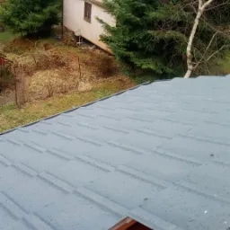 Nowe szare blaszane pokrycie dachowe z widocznymi kroplami deszczu, fragment konstrukcji drewnianej i czerwonej membrany dachowej w dolnej części kadru, w tle budynek i drzewa.