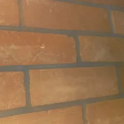 Fragment ściany z cegły klinkierowej w odcieniach pomarańczy, widoczne szare fugi, perspektywa z narożnika pomieszczenia, oświetlenie z góry.