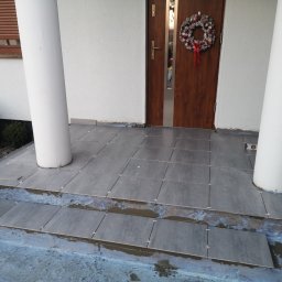 Robert chłodziński renbud - Kładzenie szarych płytek na schodach wejściowych do domu z białymi kolumnami, widoczny poziomica i świąteczny wieniec na drzwiach.