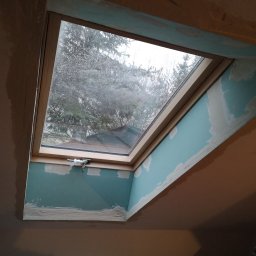 Robert chłodziński renbud - Montaż okna dachowego z widocznymi śladami szpachlowania na łączeniu z płytami, widok z dołu na tle pochmurnego nieba.