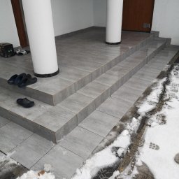 Robert chłodziński renbud - Wyłożone szarymi płytkami schody wejściowe z dwoma białymi kolumnami, na stopniach leżą czarne klapki, a obok schodów śnieg z odciskami butów.