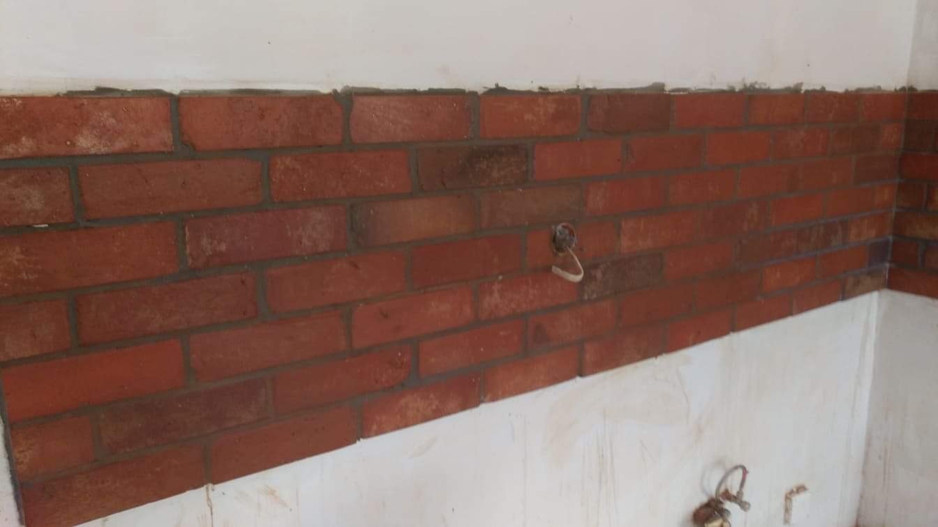 Fragment ściany z czerwonej cegły klinkierowej w trakcie układania, widoczne ślady zaprawy i niedokończone wykończenie ściany, przewód elektryczny wystający ze ściany.