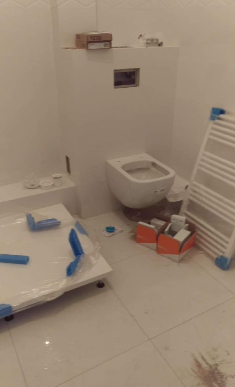 Wnętrze łazienki w trakcie remontu: podwieszana toaleta bez deski, grzejnik drabinkowy i elementy zabudowy stelaża pod umywalkę, wszystko w bieli, z widocznymi materiałami instalacyjnymi i kartonem...