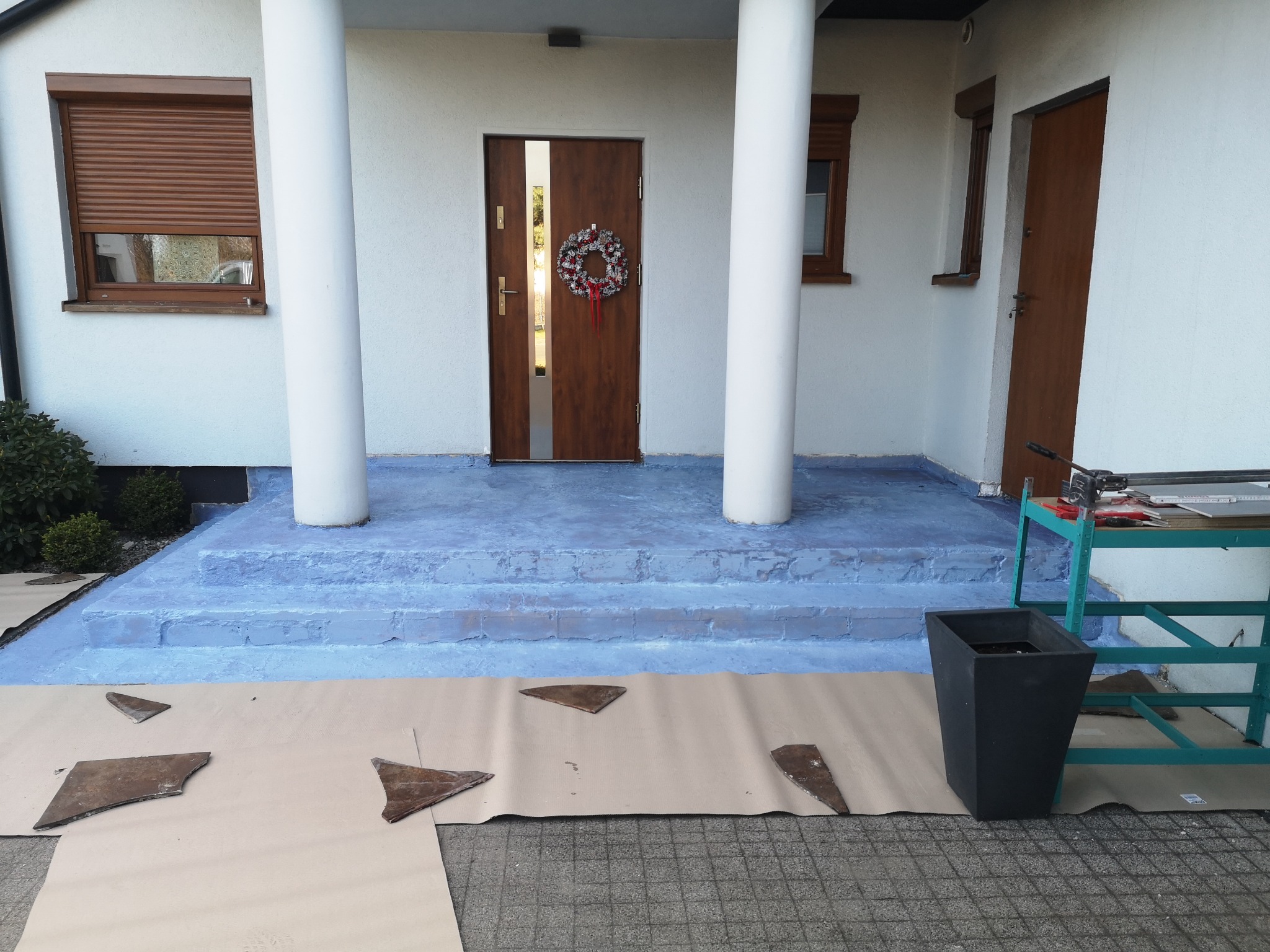 Wejście do domu z kolumnami w trakcie prac wykończeniowych: fragmenty płytek na podłożu ochronnym, pomalowane schody wejściowe na niebiesko, drzwi wejściowe z wieńcem.