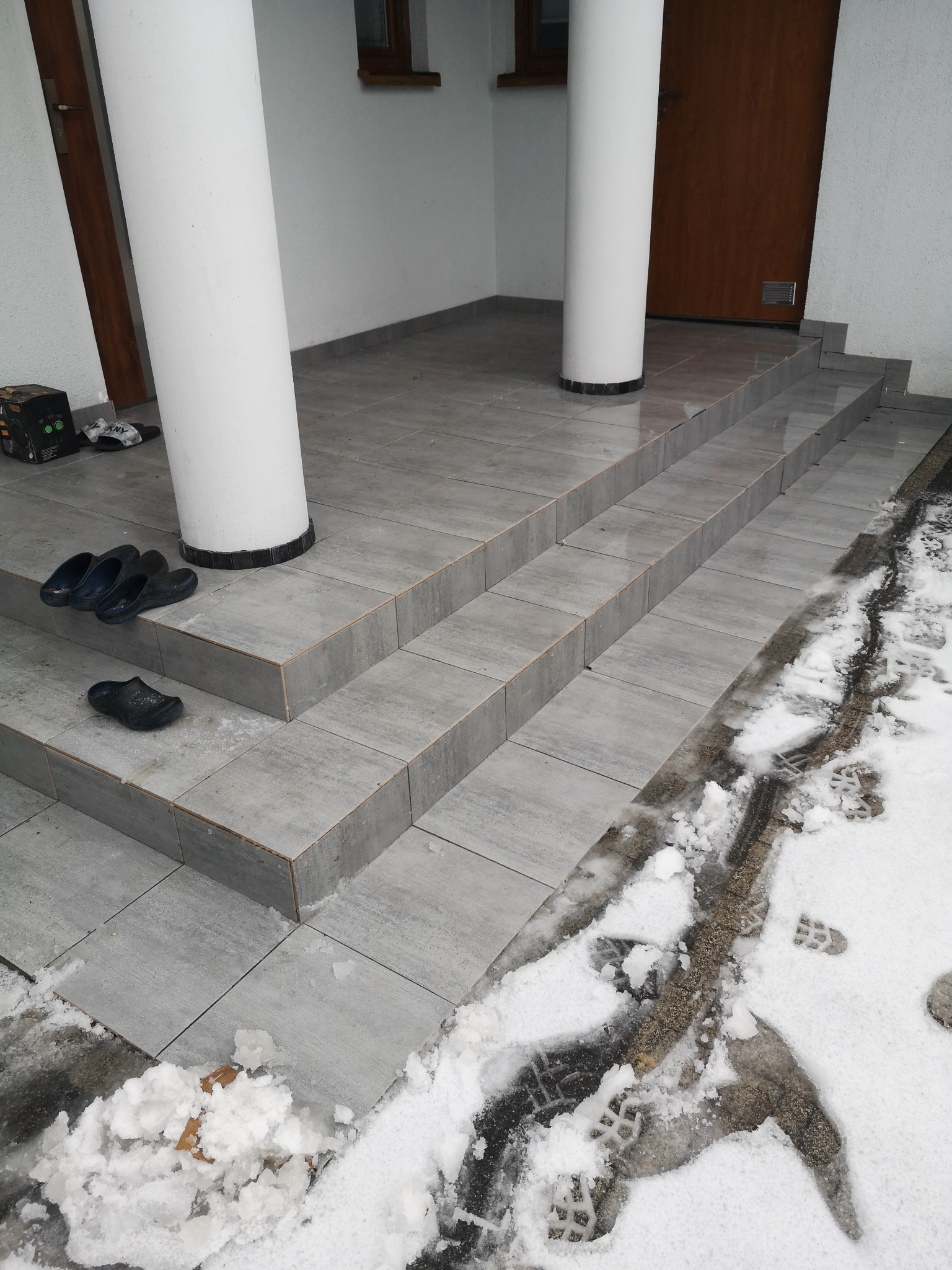 Wyłożone szarymi płytkami schody wejściowe z dwoma białymi kolumnami, na stopniach leżą czarne klapki, a obok schodów śnieg z odciskami butów.