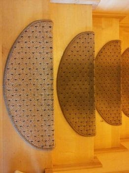 Półokrągłe wycieraczki na schodach z jasnego drewna, widoczne trzy stopnie z wycieraczkami o wzorze plecionki w odcieniach beżu i brązu.