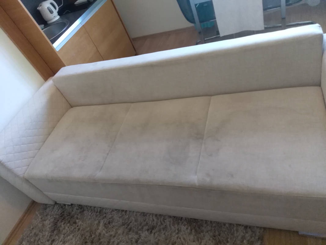 Jasna, trzysiedziskowa kanapa z widocznymi plamami na siedzisku, sfotografowana w pomieszczeniu z elementami kuchennymi w tle.