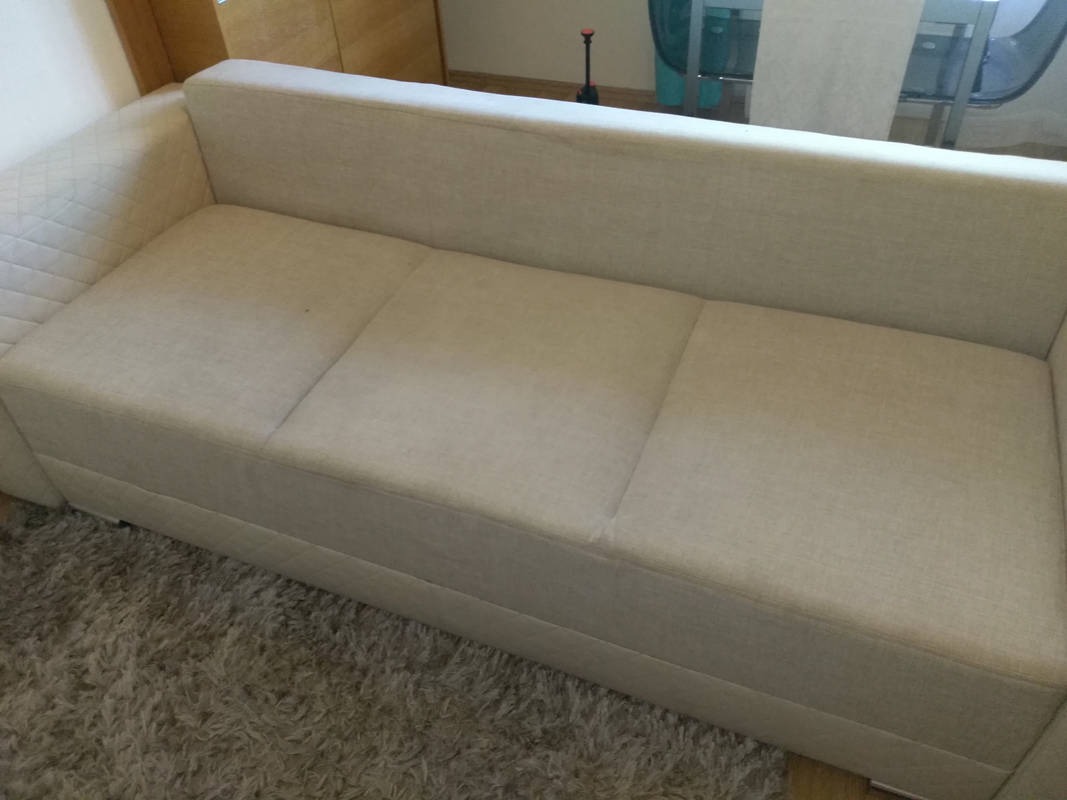 Jasna, trzyosobowa kanapa z podłokietnikiem o pikowanej fakturze, stojąca na puszystym, szarym dywanie.