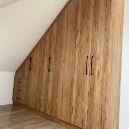 Szafa z płyty laminowanej w kolorze jasnego drewna, dopasowana do skosu poddasza, z czarnymi, podłużnymi uchwytami i trzema szufladami po lewej stronie.